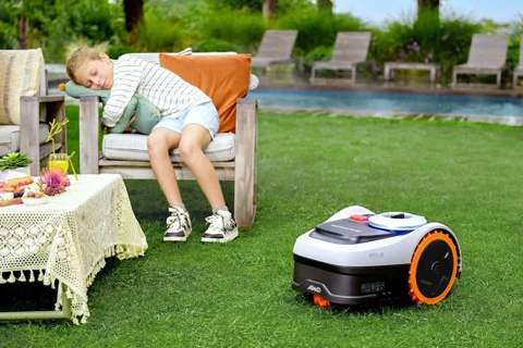 The Segway Navimow i2 AWD mower sits quietly on a lush lawn while a child sleeps in a chair nearby.
