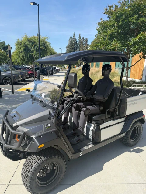 Two Figure humanoid robots sitting in a John Deere Gator utility vehicle