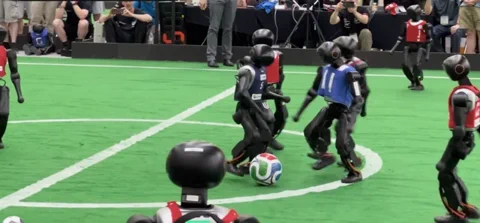 Two teams of humanoid robots playing soccer on a green field during the first 11-vs-11 match at RoboCup 2026.
